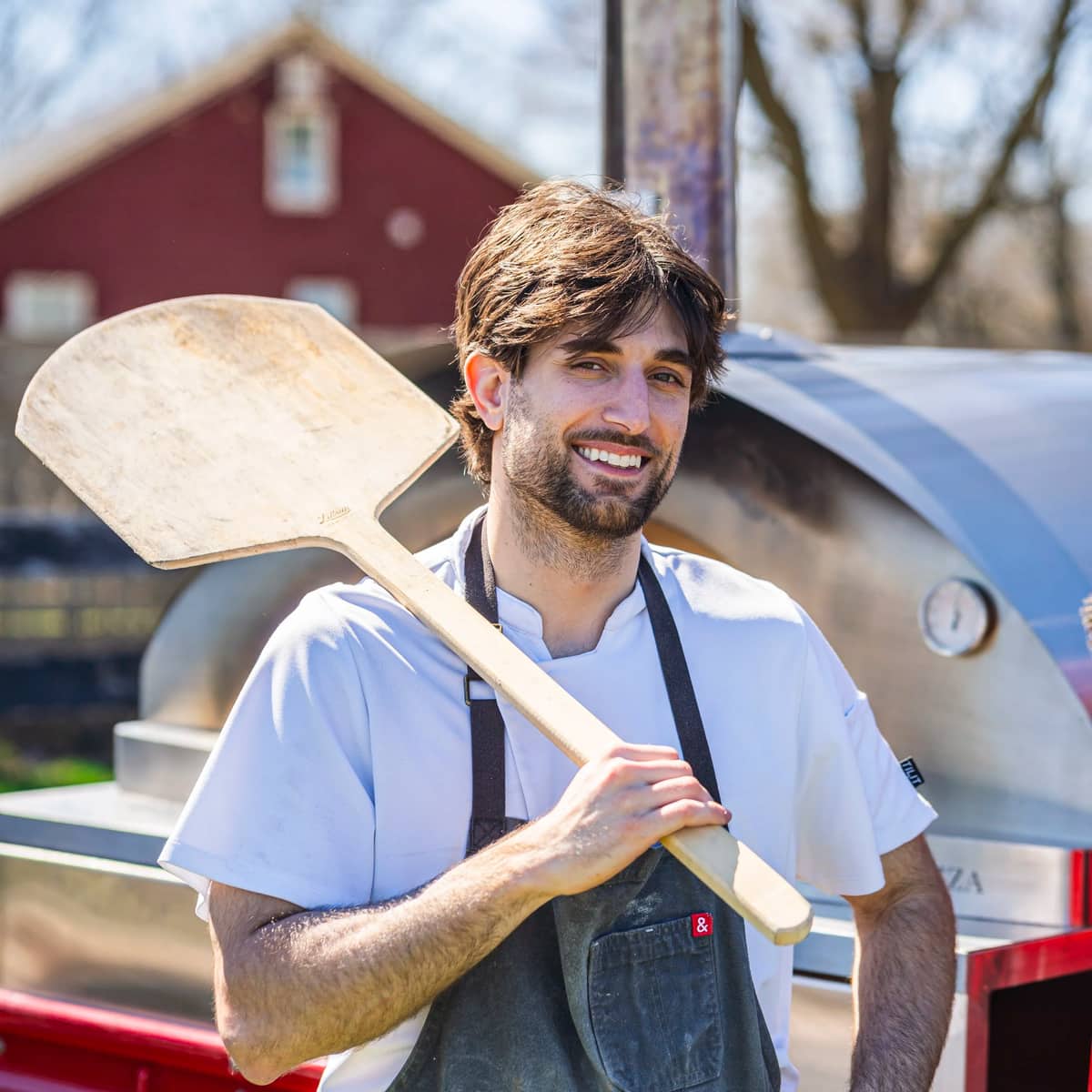 Mikey Maccarrone with a wooden pizza peel over his shoulder beside the truck's wood-fired oven