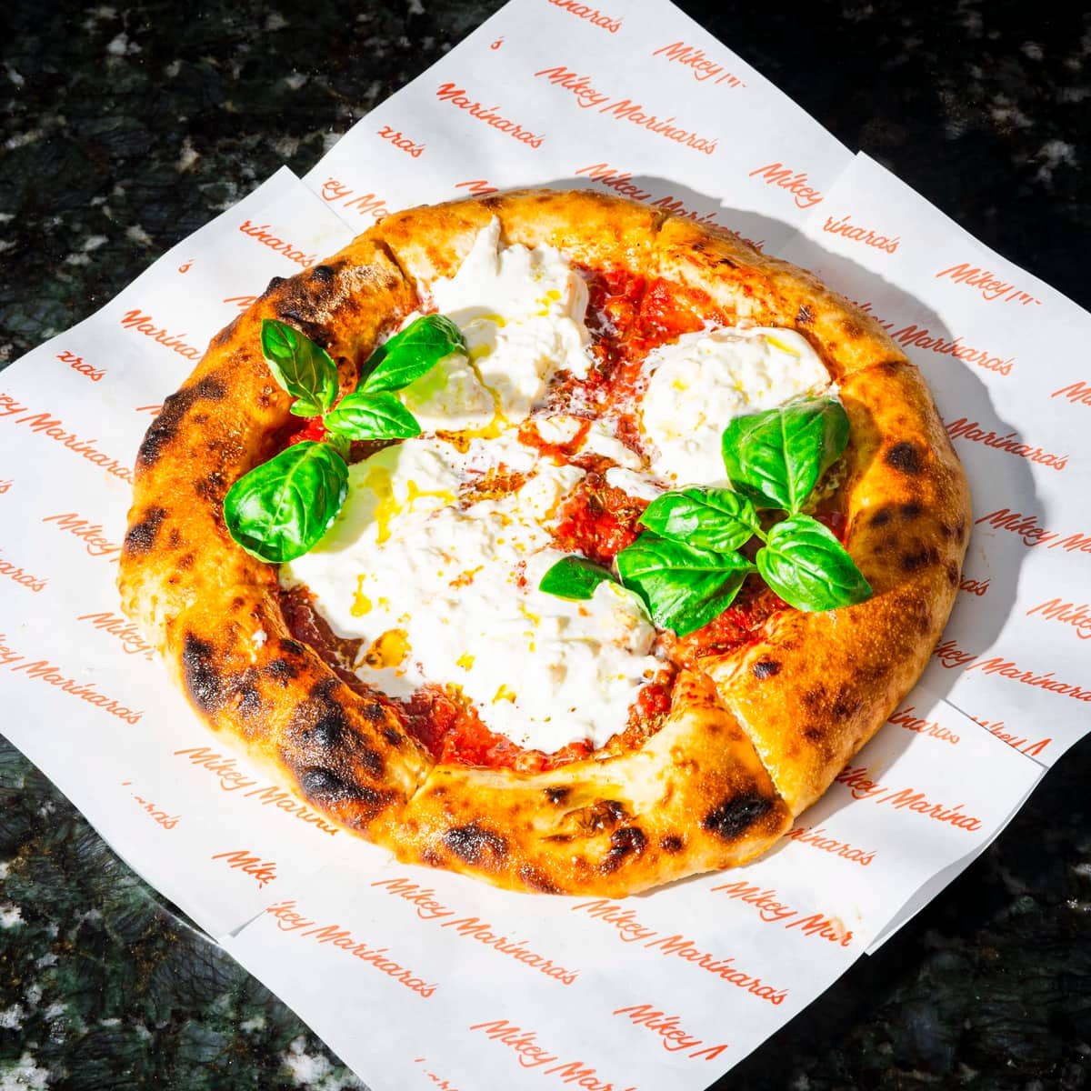 A wood-fired burrata pizza with basil on branded Mikey Marinara's paper