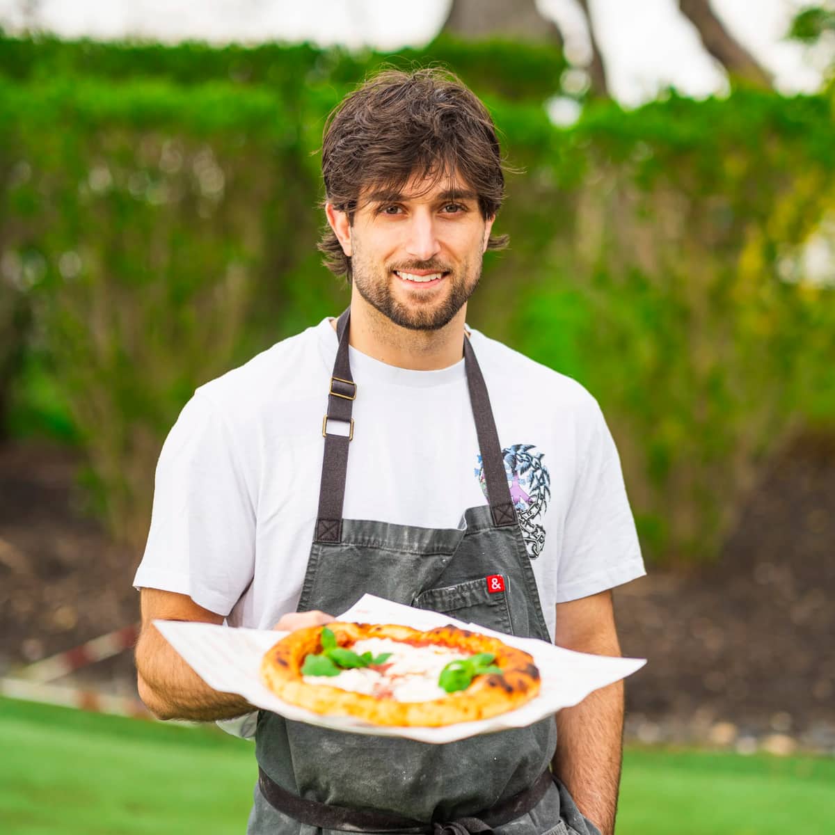 Mikey holding a finished wood-fired pizza on a lawn