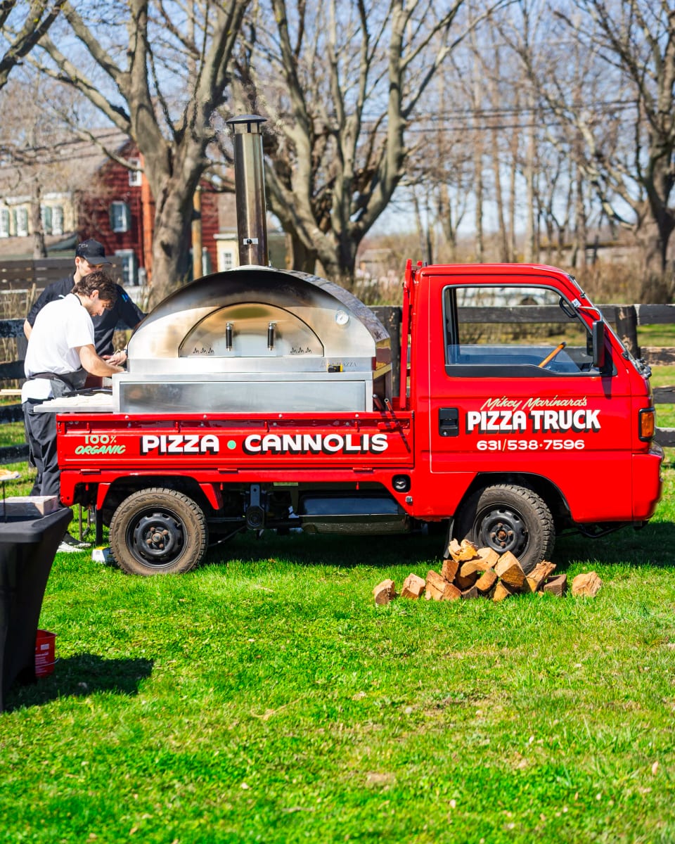 The red pizza truck on a back lawn with its wood-fired oven lit and split firewood stacked beside the wheel